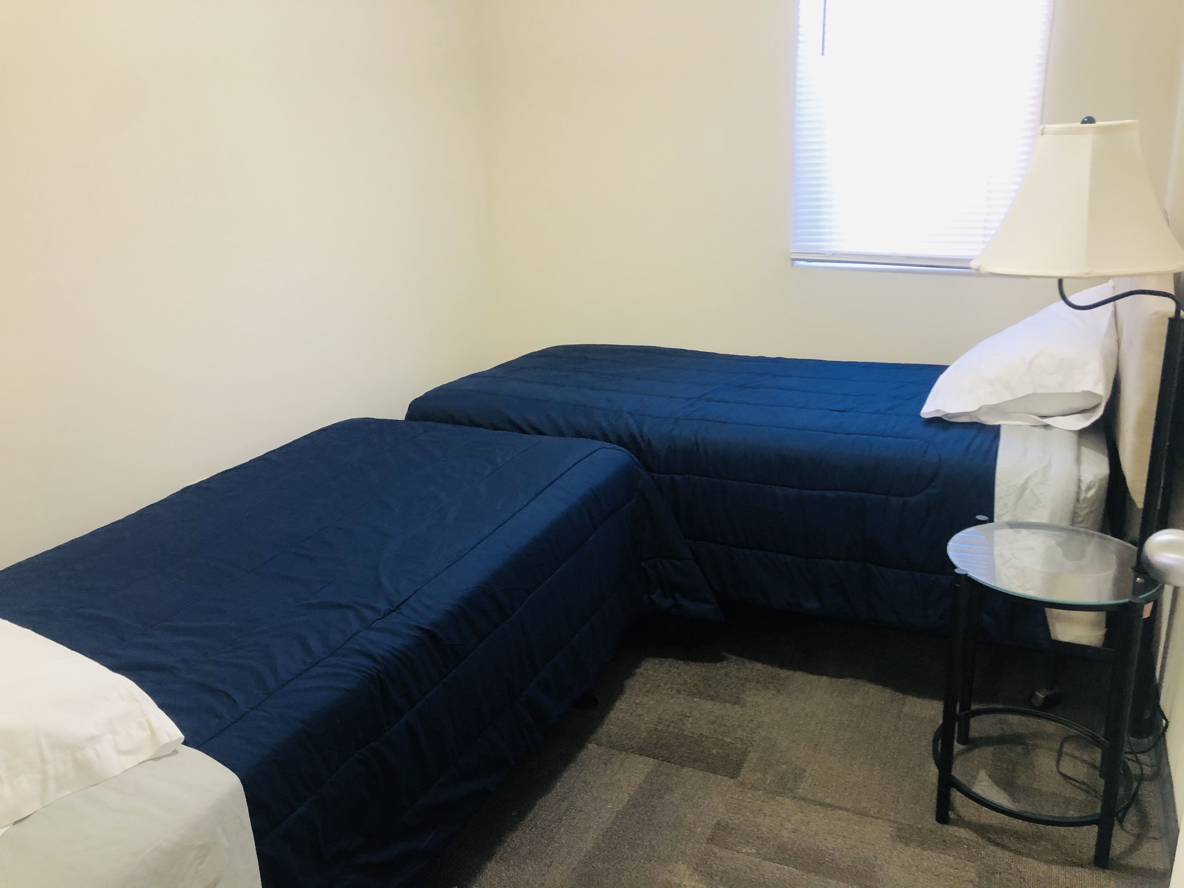 A small room with two beds, a nightstand, a lamp, and a window with blinds.