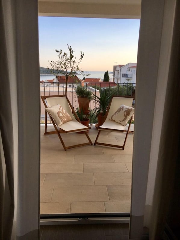 A balcony with two wooden chairs and a potted plant, overlooking a coastal area with buildings.
