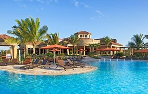 A pool area with lounge chairs and umbrellas, palm trees, and a building with a tiled roof. - Lovely 2-Bedroom in Naples' highly desirable Treviso Bay community (Naples)