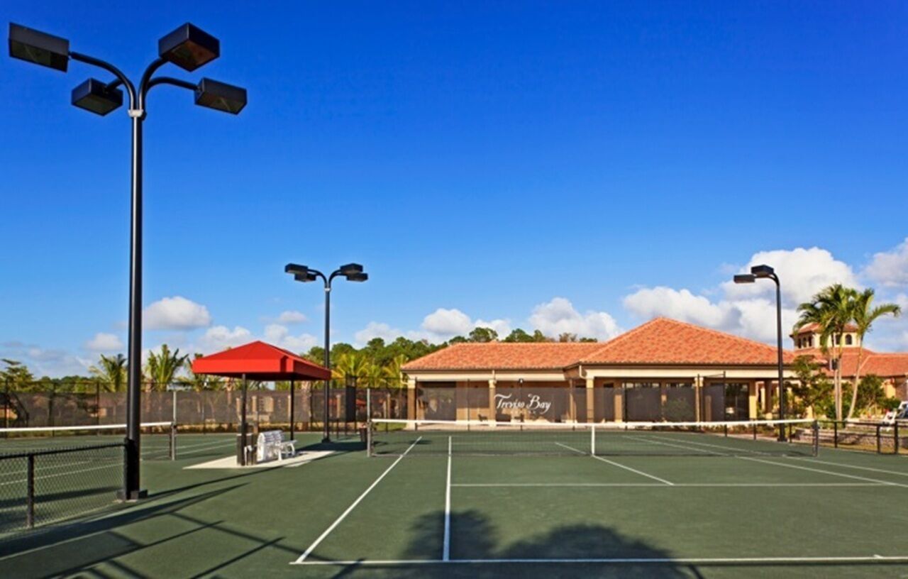 An outdoor tennis court with multiple light poles, a covered seating area, and a building with a red roof in the background.