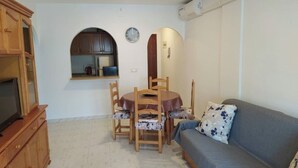 A compact living space with a dining table, chairs, a sofa, a television on a wooden cabinet, and a kitchen area visible through an archway. - Everything ready to enjoy the summer, swimming pools, near the sea, tennis and more... (Torrevieja)