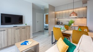 A modern living room with a flat-screen TV, a dining area with a table and chairs, and a kitchen with wooden cabinets and pendant lighting.