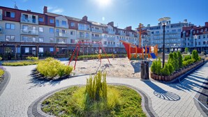 A playground with red equipment, surrounded by a paved area with plants and a bicycle lane.