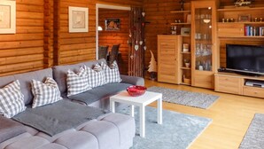 En mysig stuga med träpanel på insidan, en grå soffa, rutiga kuddar, en röd glasskål på ett vitt bord och en tv på ett träställ.