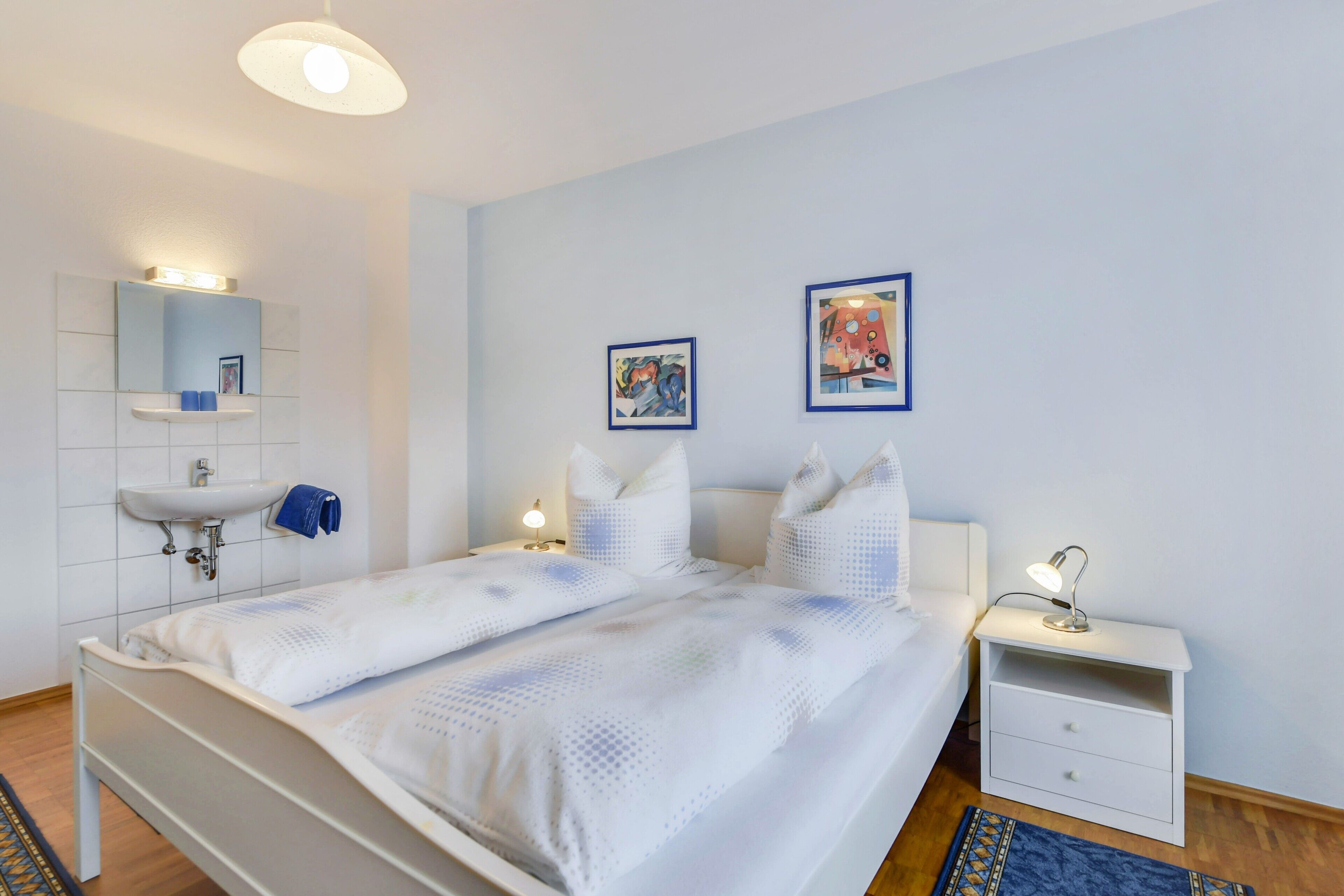 Ein Schlafzimmer mit einem Bett, Nachttischen, einem Waschtisch an der Wand und zwei gerahmten Bildern.