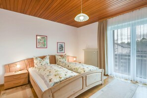 A bedroom with a wooden ceiling, two bedside tables with lamps, a bed with floral bedding, and a large window with sheer curtains.