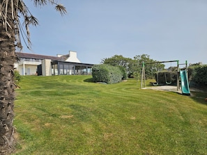Un giardino curato con prato verde, altalena e scivolo, circondato da vegetazione rigogliosa e una casa moderna con ampie finestre.