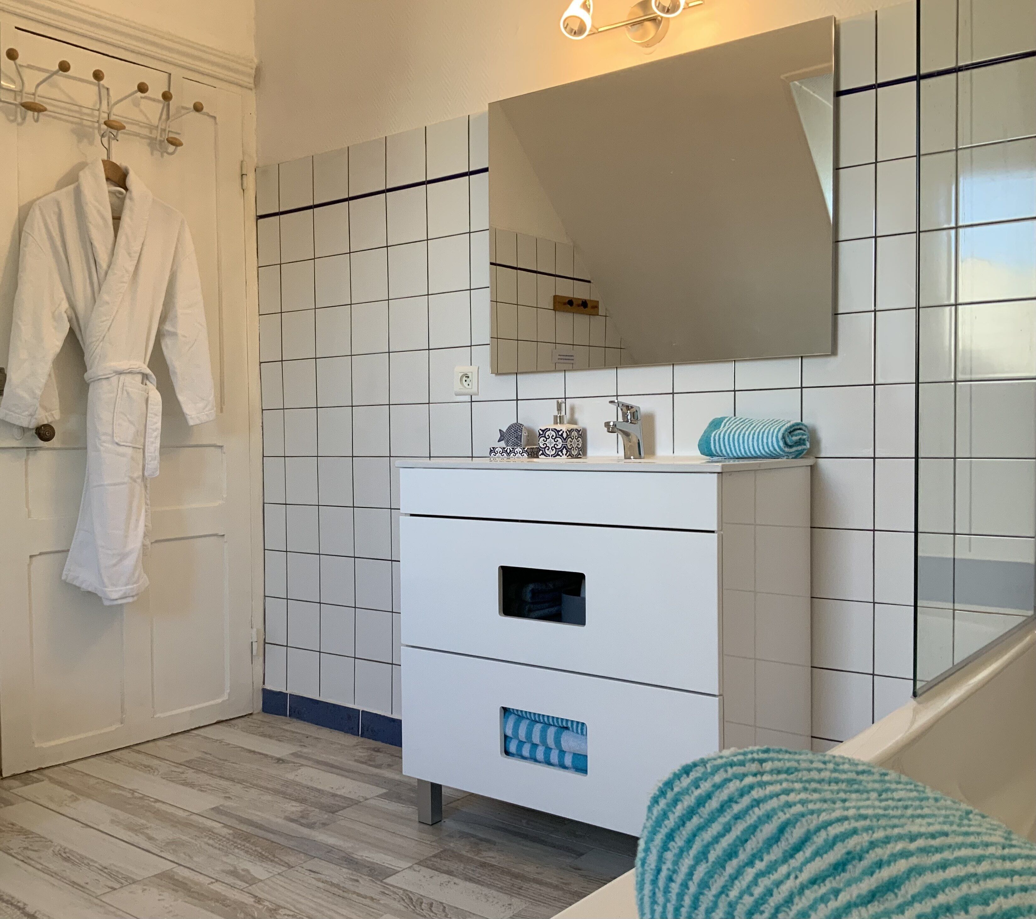 A bathroom with white tiled walls, a white sink with a mirror, and a wooden floor.