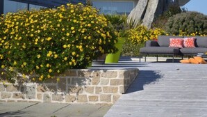 A patio area with a stone planter, a gray sofa with red and white pillows, and yellow flowers. - CHARMING APARTMENT ⭐️⭐️⭐️⭐️ WITH SWIMMING POOL BEAUTIFUL SEA VIEW 150 M FROM SANDY BEACH (SIBIRIL)