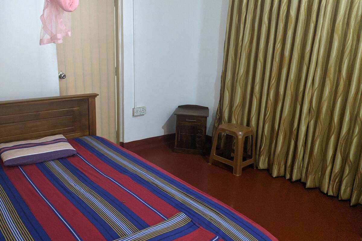 A room with a wooden bed, a striped bedspread, a small stool, a radio, and a curtain.