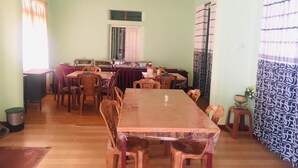 A dining area with a long wooden table and multiple chairs, a kitchen area with appliances, and a patterned curtain on the right side.