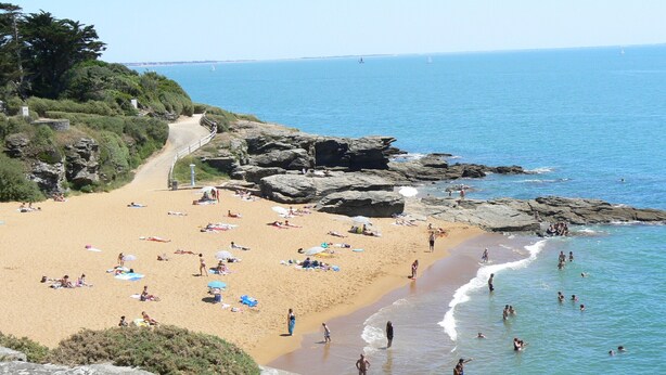 Ein Strand mit Menschen, die sonnenbaden und schwimmen, Felsformationen und freiem Blick auf das Meer.
