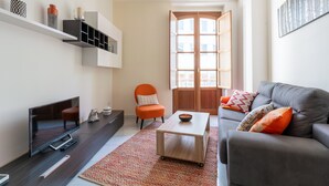 Un salon moderne avec un canapé gris, un fauteuil orange et un téléviseur posé sur un meuble en bois foncé. Une table basse en bois et un tapis à motifs se trouvent au centre de la pièce.
