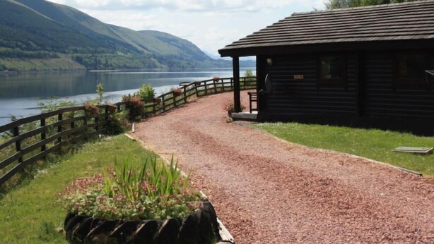 A wooden cabin with a gravel path leading to it, surrounded by greenery and a lake. - Braeside by Interhome (Invergarry)
