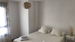 Ein ordentlich bezogenes Bett mit weißen Leinen und Kissen, ein Nachttisch mit einer Vase getrockneter Blumen und eine gewebte Pendelleuchte.