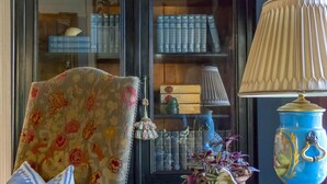 Ein Zimmer mit einem dunklen Holzschrank mit Glastüren, einem blumigen Sessel, einer blauen Lampe und einer gemusterten Bettdecke.