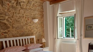 Un dormitorio con pared de piedra, techo de madera, cama con cabecera blanca, ventana con cortinas y radiador.