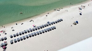 An aerial view of a beach with rows of umbrellas and beach chairs, a clear blue ocean, and a sandy shore.