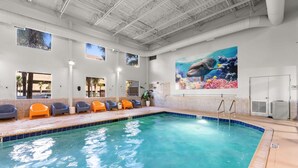 An indoor swimming pool with clear blue water, a mural of a hand holding a fish, and several orange and gray lounge chairs along the poolside.