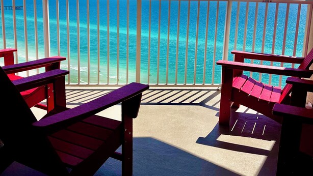 A balcony with a view of the ocean, featuring two Adirondack chairs and a railing.