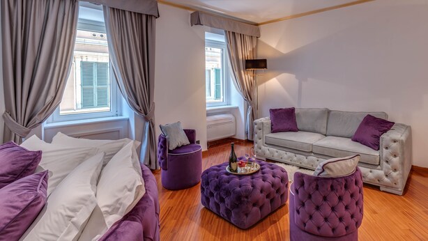 A modern room with a bed, a grey sofa, and a purple ottoman. There are curtains on the windows, and a floor lamp is visible. - SPANISH STEPS Extra Luxury Apartment (Roma)