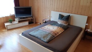 A modern bedroom with a wooden floor, a bed with a patterned bedspread, a television on a wooden stand, and a small plant on the nightstand. - Apartment on the farm (Gadmen)