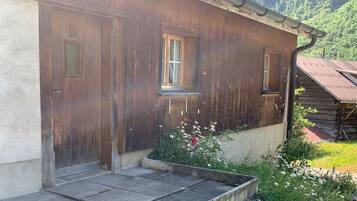 Un bâtiment rustique en bois, doté de deux portes et de fenêtres, avec une marche en béton menant à l’entrée, et un jardin planté de fleurs sauvages.