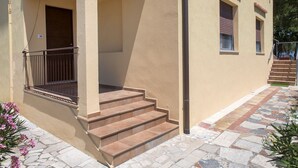 Un bâtiment à la façade beige, doté d’une entrée couverte avec une rambarde en métal et d’un escalier en pierre menant à une porte.