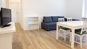 Un salon moderne avec un téléviseur à écran plat, un canapé gris, une table à manger blanche avec des chaises et un plancher en bois.