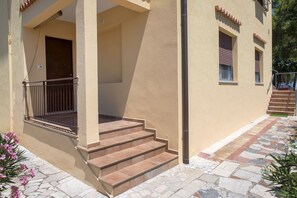 Un edificio con fachada color beige, un acceso cubierto con barandilla metálica y un conjunto de escalones de piedra que conducen a una puerta.