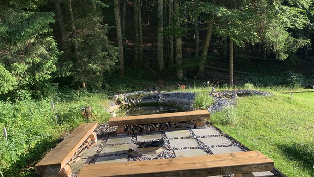 Un área de estar al aire libre de estilo rústico con suelo de baldosas de piedra y bancos de madera, rodeada por un entorno de bosque natural.