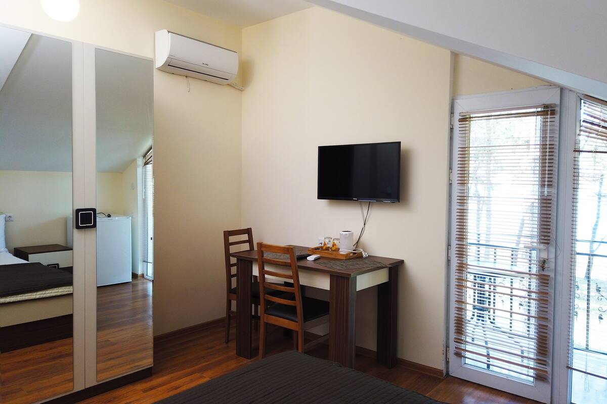 A compact room with a bed, a desk with a chair, a flat-screen TV, and a window with blinds.