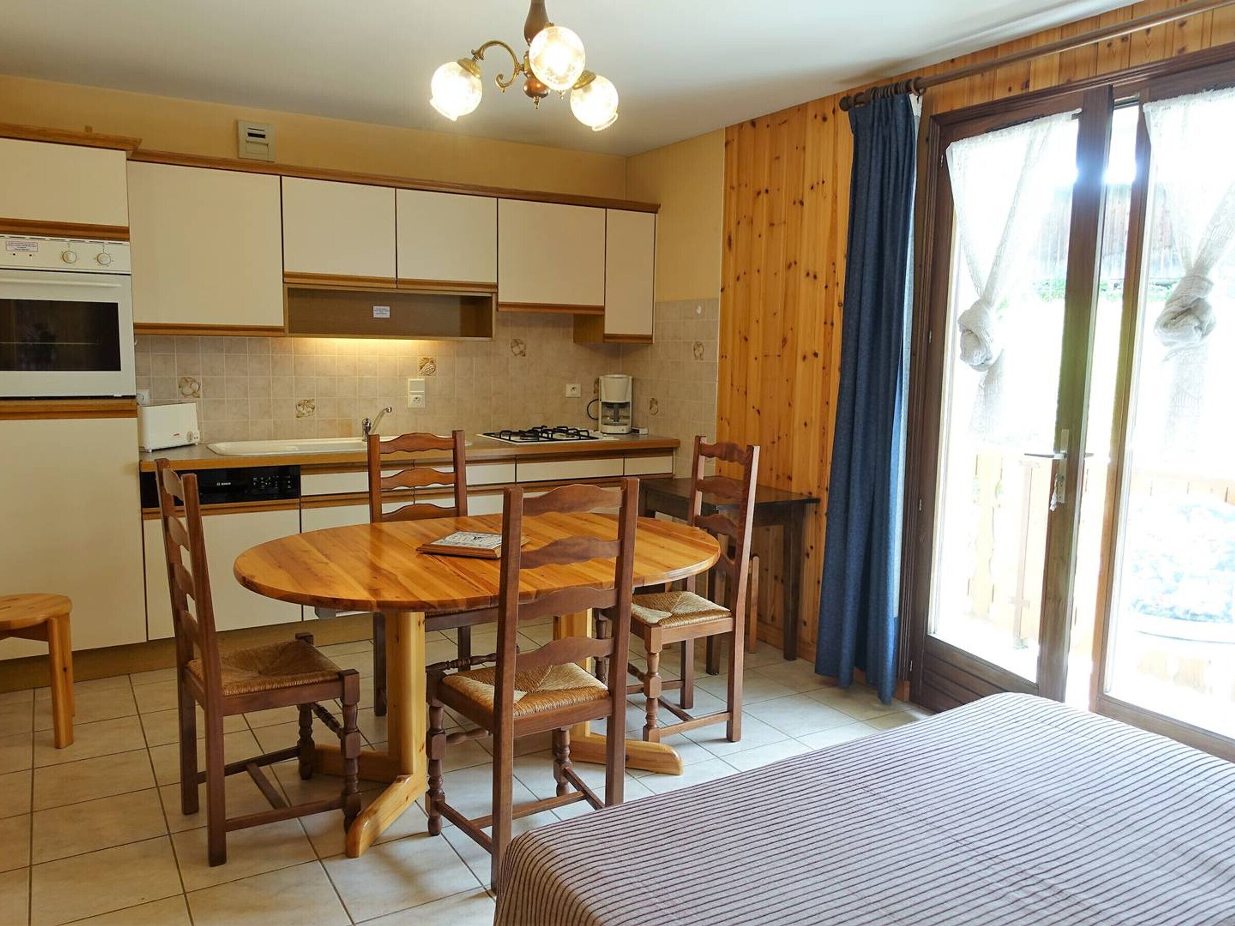 A compact kitchen with a dining area, wooden cabinets, a stove, and a large window with curtains.