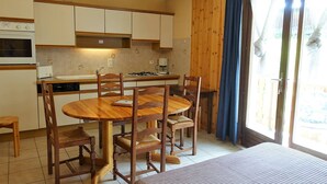 A compact kitchen with a dining area, wooden cabinets, a stove, and a large window with curtains. - Quiet apartment for 4, at the foot of the slopes with parking (Arêches-Beaufort)