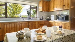 A kitchen with wooden cabinets, a window with a view of trees, a table with a floral tablecloth, and various breakfast items.
