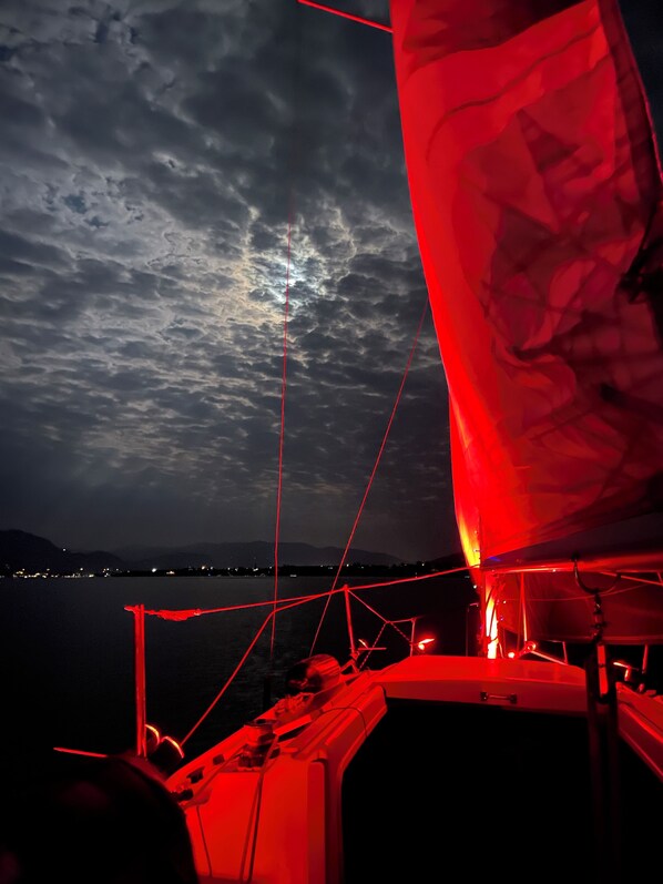 A boat with a red sail and a person at the helm, set against a backdrop of a cloudy sky and distant mountains.