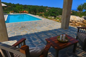 A pool area with stone flooring, outdoor furniture, and a view of the sea and hills.