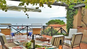 Un área de comedor al aire libre con vistas al mar, mesas dispuestas con platos y vasos, y una botella de vino.