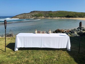 A table set up on a grassy area with a view of the ocean and a rocky hill in the background. - Romantic Beachfront Paradise Villa with Incredible Ocean Views & Outdoor Event Space (Islote Arecibo)