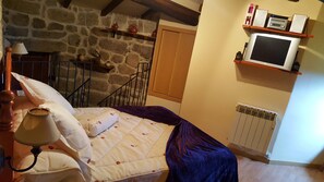 A bedroom with a stone wall, a bed with a white and purple bedspread, a wooden headboard, a bedside lamp, a shelf with a radio and books, and a window with a wooden shutter.