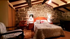 Um quarto aconchegante com parede de pedra, estrutura de cama de madeira, mesa de cabeceira, uma cadeira e uma pequena mesa com abajur.