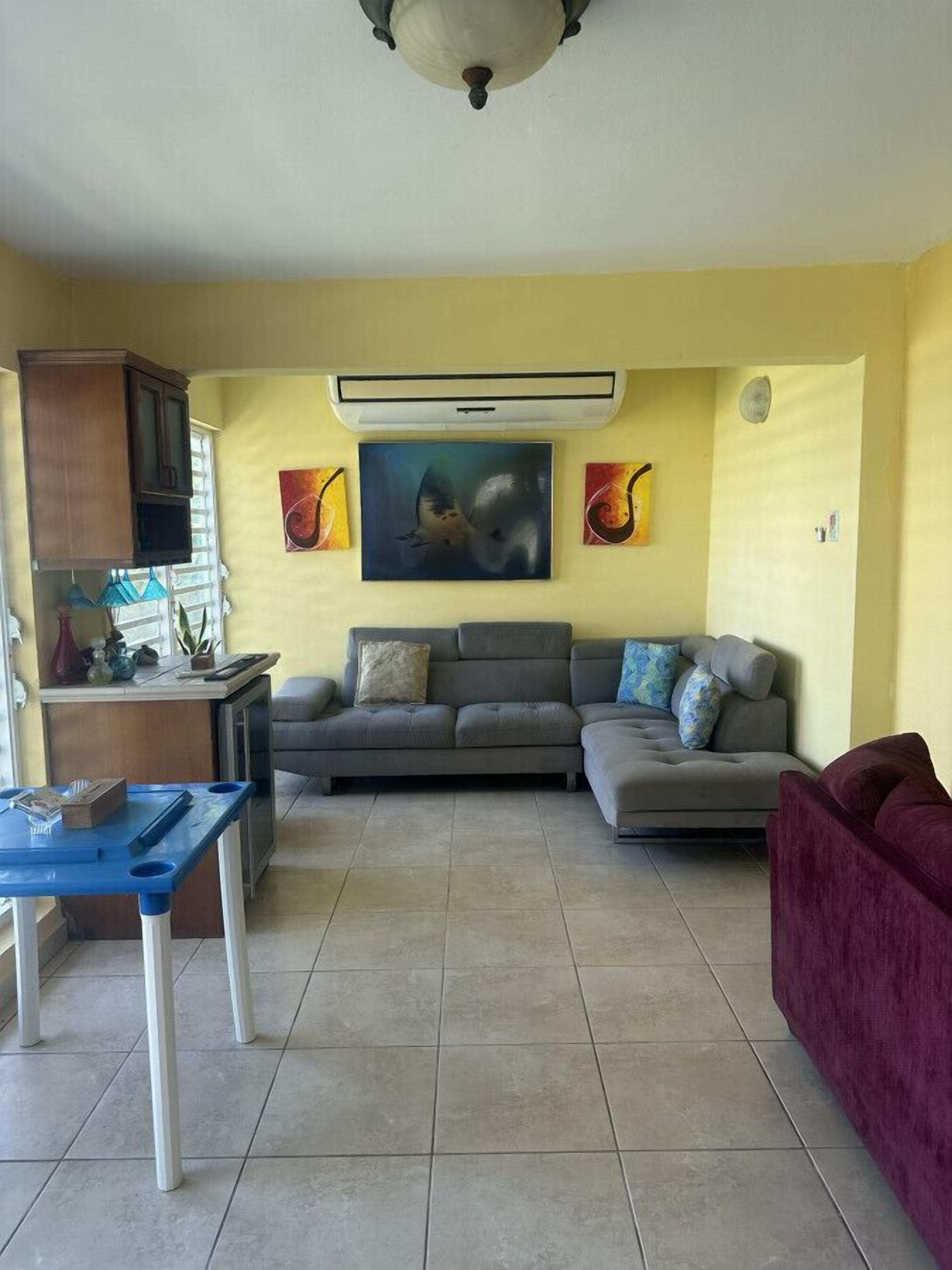 A living room with a sofa, a red armchair, a blue table, and a TV. There are paintings on the wall and an air conditioning unit above the sofa.