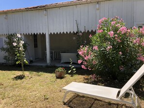 A white building with a covered patio area, a white chair, and pink flowering plants.