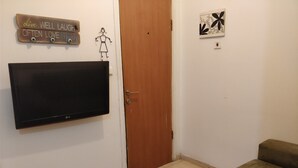 A living room with a flat-screen TV mounted on the wall, a wooden door, a small artwork on the wall, and a brown couch. - Cozy 2 Bedroom Apartment by The Beach (Tel Aviv)