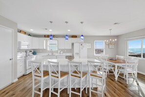 A modern kitchen with white cabinets, a central island, and a dining area with white chairs. - Beachfront Cottage | Covered Deck | West Beach Location (Gulf Shores)