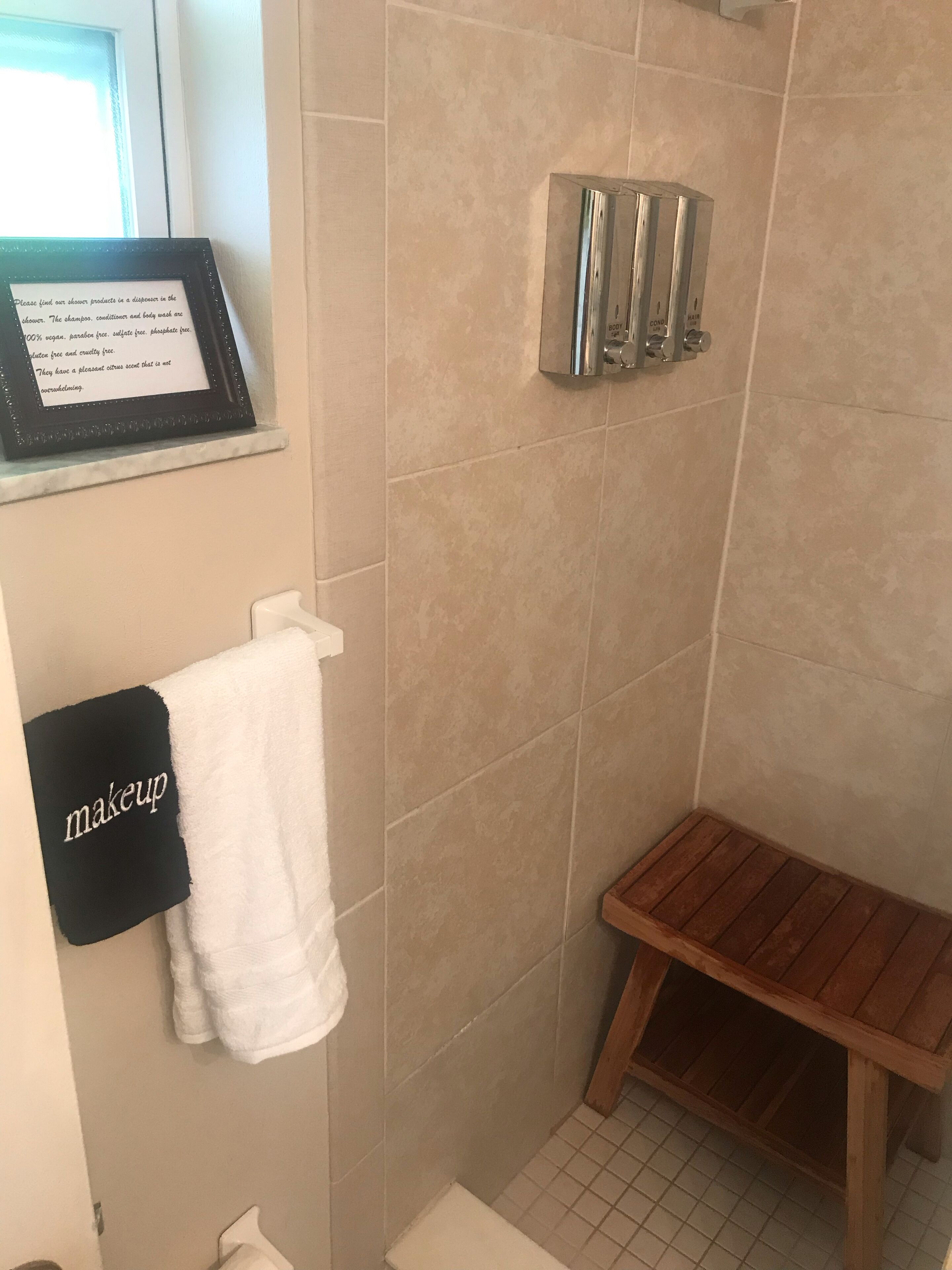 Een badkamer met een douche, een lijst met een bordje, een handdoekhouder met een handdoek voor make-up, en een houten bankje.