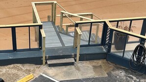 Un escalier en bois menant à une plage, avec une rambarde, une canne à pêche et un récipient bleu sur le sable.