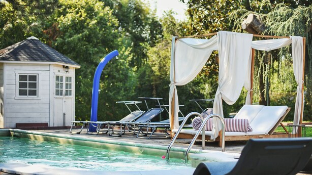 Ein Poolbereich mit Liegestühlen, einem kleinen Gebäude und einer Sonnensegelkonstruktion.