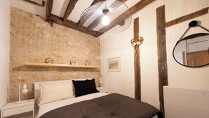 Un dormitorio con pared de piedra, una cama con cubrecama gris oscuro, una mesita de noche blanca con lámpara y un estante de madera con máscaras.