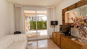 A modern living room with a white sofa, a wooden TV stand, and a glass door leading to a balcony with a view of the city.